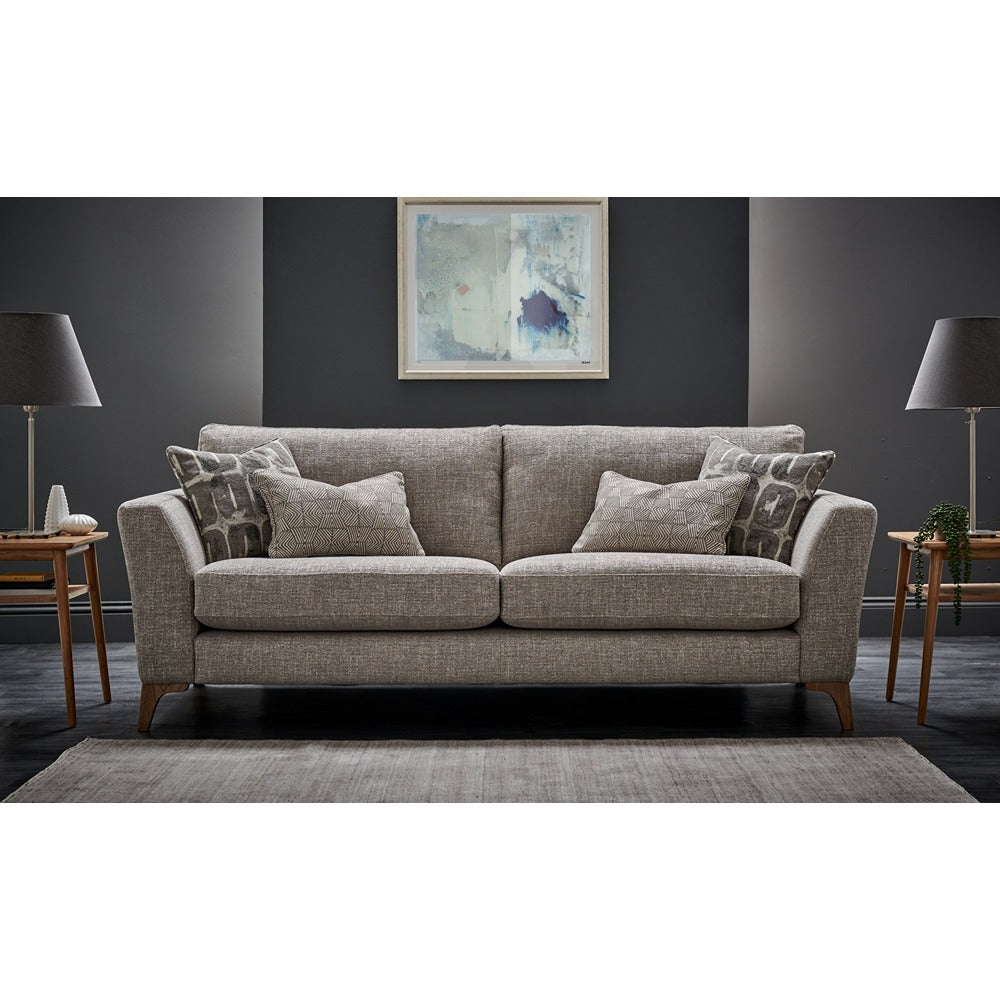 A modern two-seater sofa in textured beige fabric, styled with four matching throw pillows and two patterned accent pillows, set in a contemporary living room with dark grey walls, wooden side tables, lamps, and a rug.