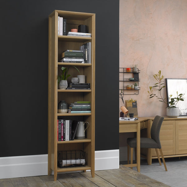 Tall wooden bookshelf against a black wall with a home office setup in the background.