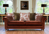 A classic brown velvet sofa sofa in a beautifully decorated room with patterned back scatter cushions, side tables, lamps, and large windows.