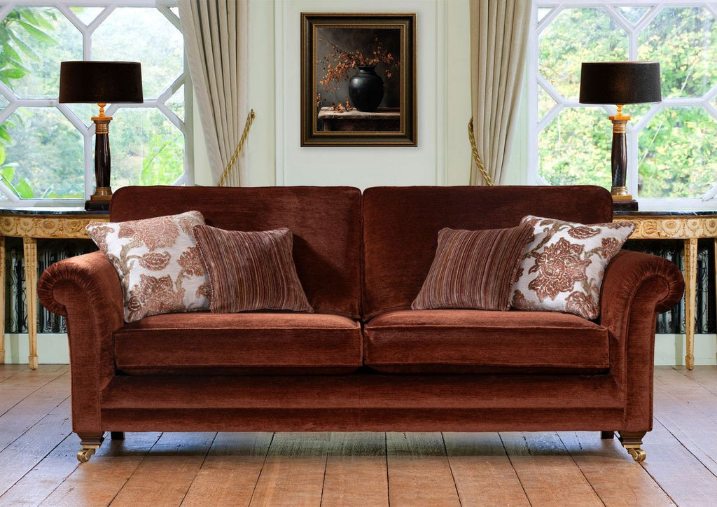A brown velvet three seater sofa with rolled arms and gold-toned legs, styled with four decorative throw pillows in a bright room with large windows, lamps, and a framed painting.