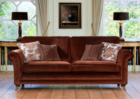 A brown velvet three seater sofa with rolled arms and gold-toned legs, styled with four decorative throw pillows in a bright room with large windows, lamps, and a framed painting.