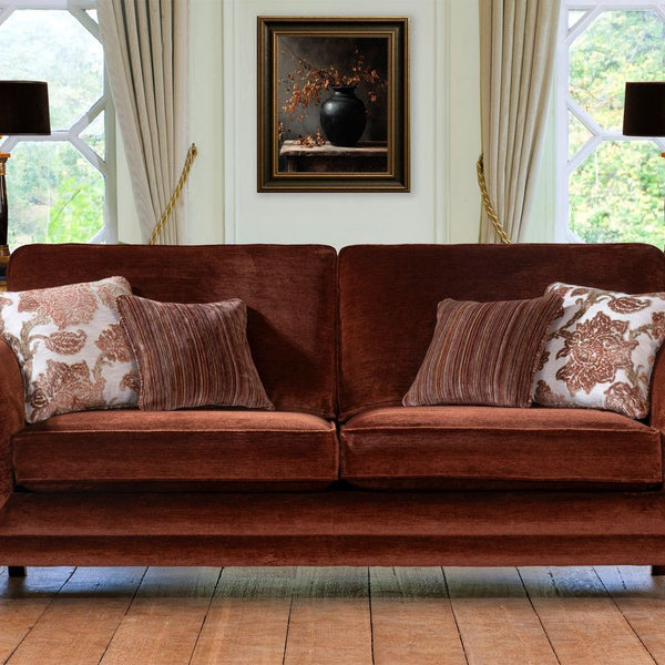 A brown velvet three seater sofa with rolled arms and gold-toned legs, styled with four decorative throw pillows in a bright room with large windows, lamps, and a framed painting.