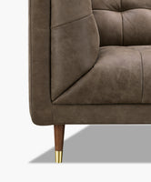 Close-up of a taupe brown leather upholstered armchair showing the tufted backrest, seat cushion, and a wooden leg with a gold metal cap on a white background.