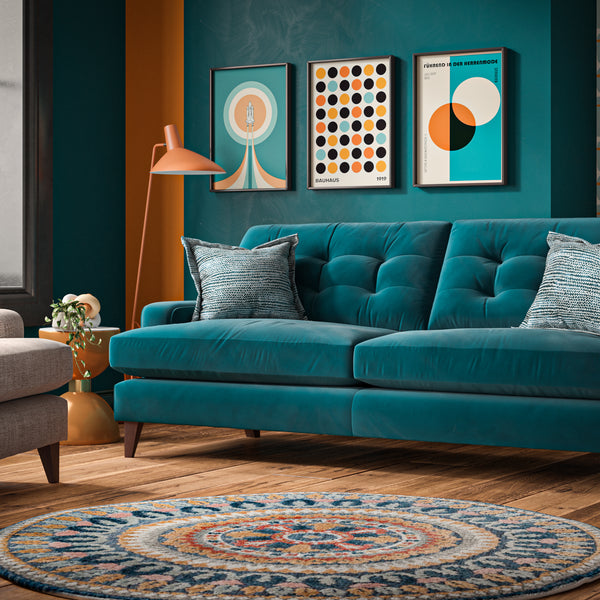 A teal velvet tufted sofa in a modern living room setting, styled with two blue-grey scatter cushions, a round patterned rug, a grey armchair, and colourful wall art.