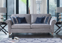 A light grey upholstered sofa with rolled arms and gold-toned legs, styled in a modern living room with blue and navy throw pillows, side tables, lamps, and large windows.
