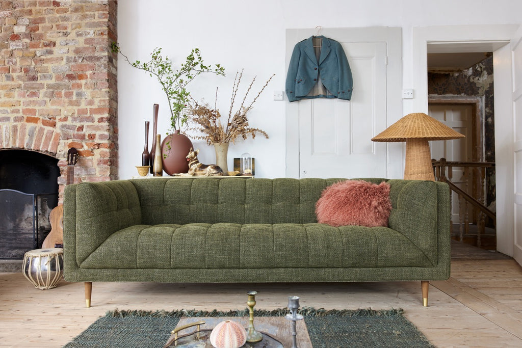 An olive green tufted fabric sofa in a styled living room setting, featuring a pink shaggy throw pillow, a brick fireplace on the left, and a woven floor lamp on the right.