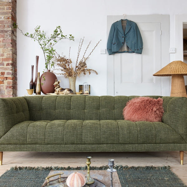 An olive green tufted fabric sofa in a styled living room setting, featuring a pink shaggy throw pillow, a brick fireplace on the left, and a woven floor lamp on the right.