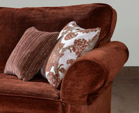 A close-up of a brown velvet sofa with two decorative scatter cushions, one solid brown and one white with a brown floral pattern, set against a neutral background.
