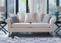 A light beige upholstered sofa with rolled arms, and castors, styled in a modern living room with grey and beige patterned scatter cushions, next to black side tables with silver lamps.