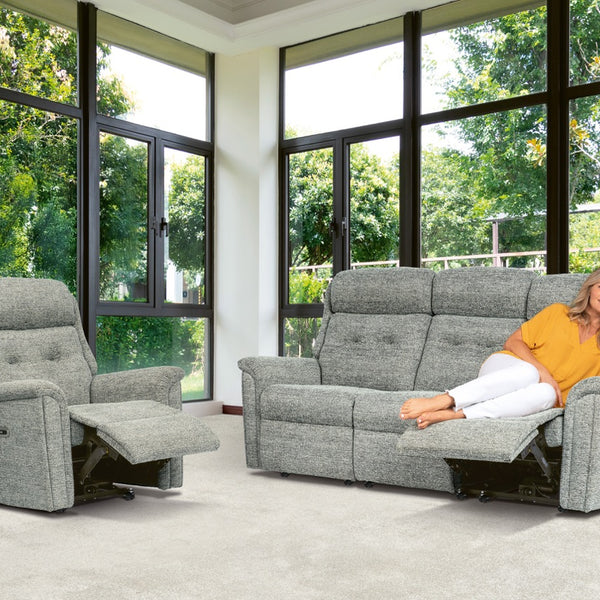 Woman relaxing on a grey reclining sofa in a room with large windows and greenery outside.