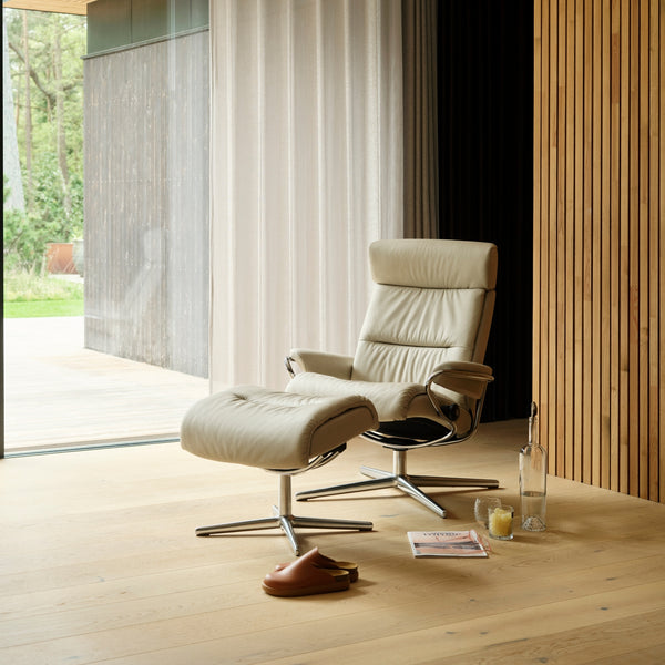 Modern beige recliner chair with stool in a room with wooden floor and large windows.