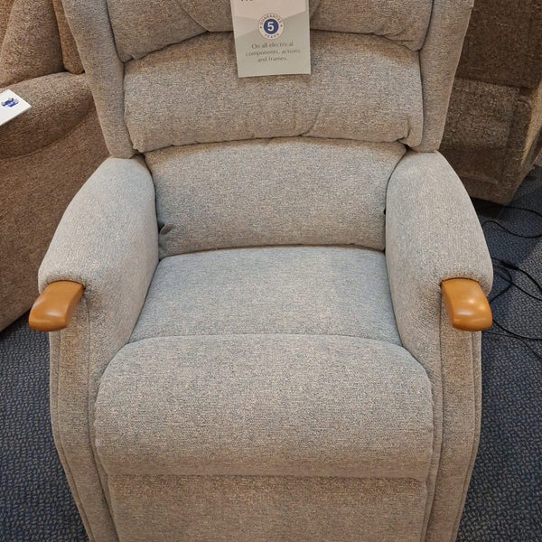A light beige textured fabric recliner chair with wooden armrests and a white paper tag attached to the backrest, photographed in a furniture showroom.